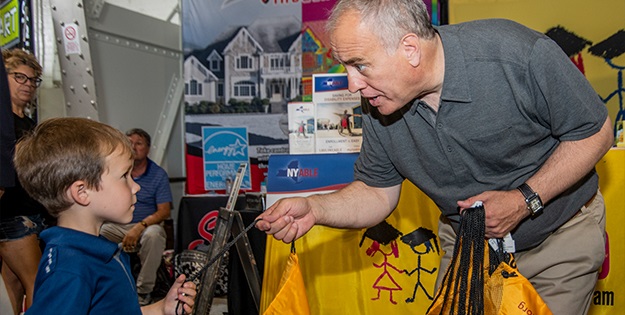 State Comptroller DiNapoli bends down to hand a 529 giveaway backpack to a young boy at the NYS Fair 