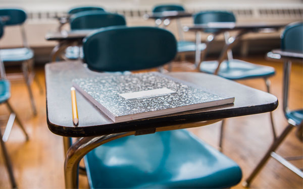 Close-up of a school desk inside a classroom with a notebook and pencil on top.  
