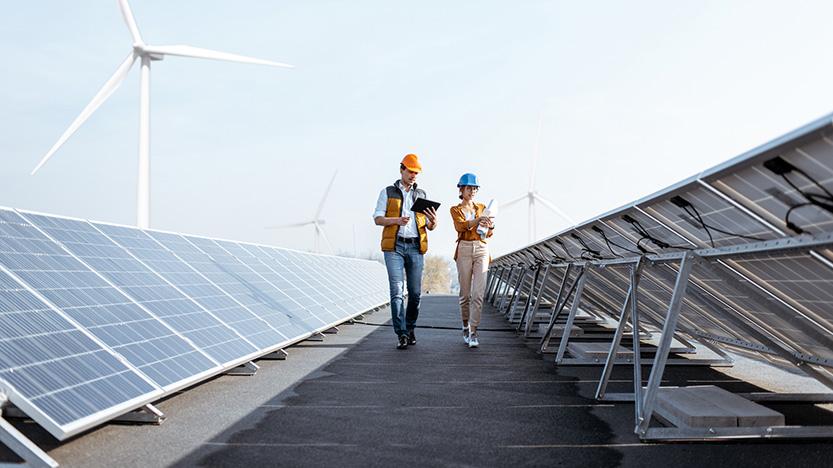 Two engineers walking and examining solar panels with a wind turbine in the background. 